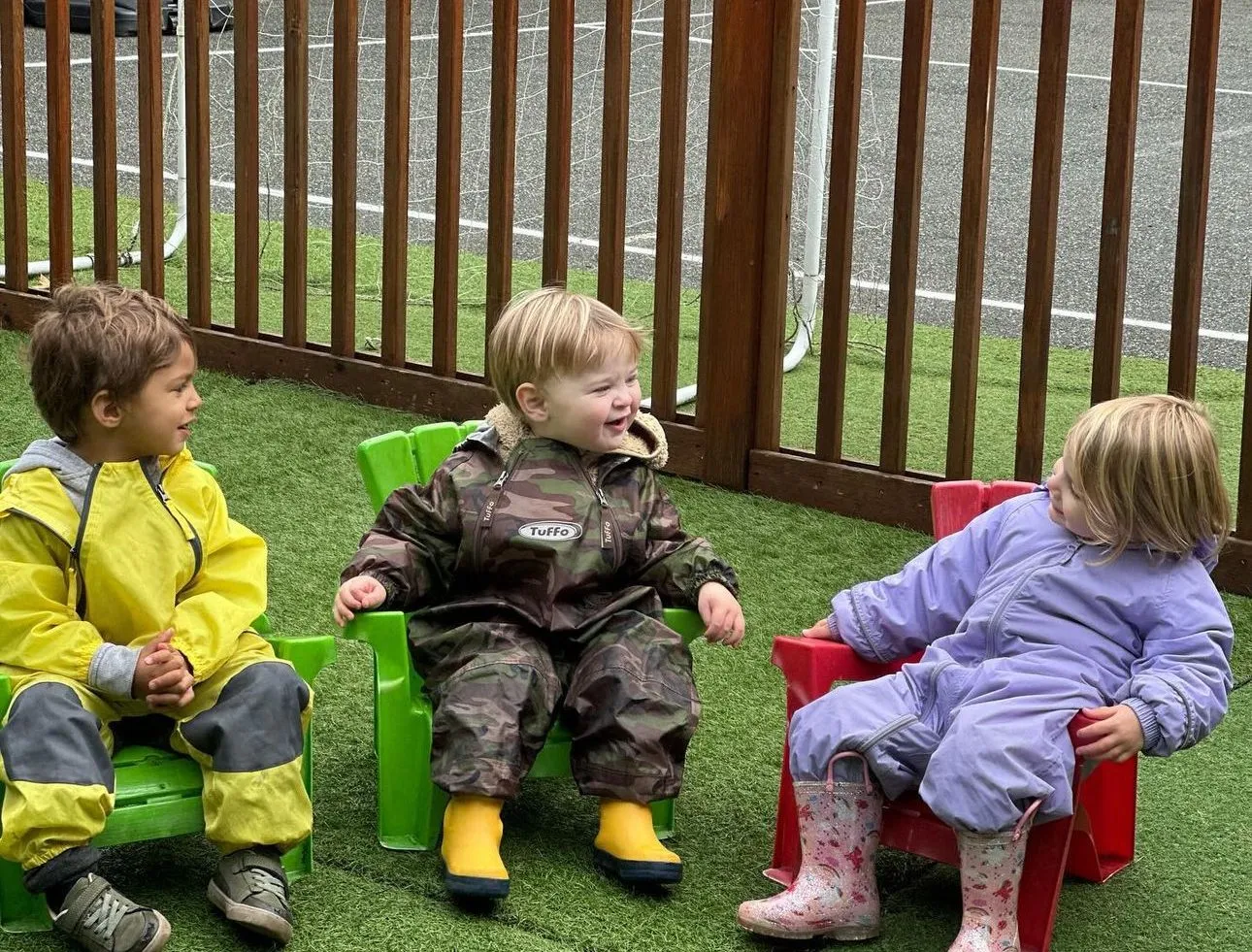 Three toddlers in rain suits and boots sitting on colourful chairs in the outdoor play yard