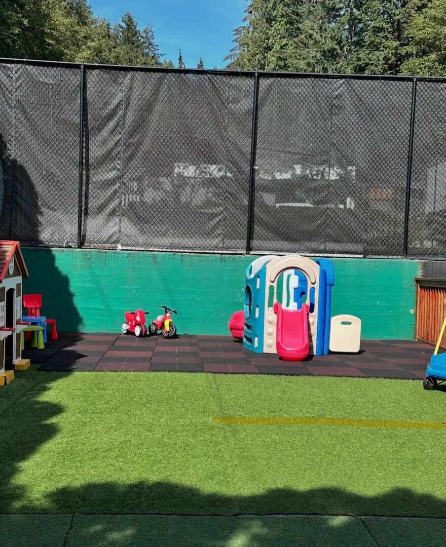 Fenced outdoor play yard with artificial turf, a slide, ride-on toys and a playhouse